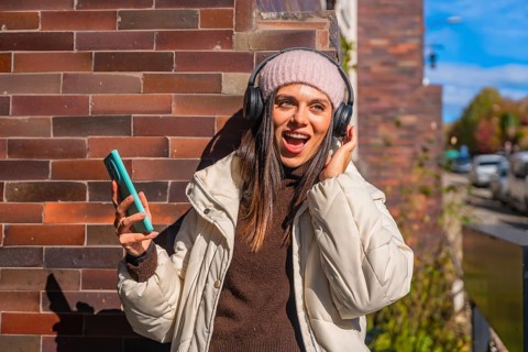 Young woman singing along to music on wireless headphones, holding her phone against a brick wall