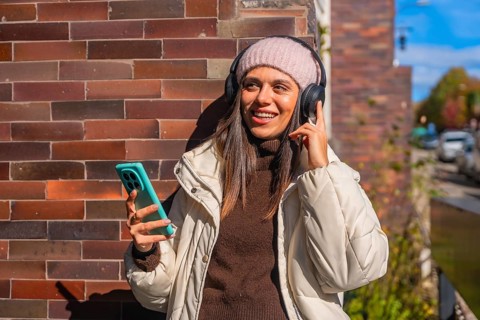 Young woman smiling while enjoying music through her headphones, holding a smartphone outdoors
