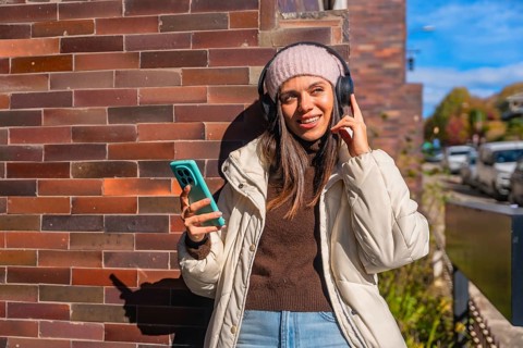 Smiling young woman dressed in warm winter clothes standing by a brick wall, peacefully listening