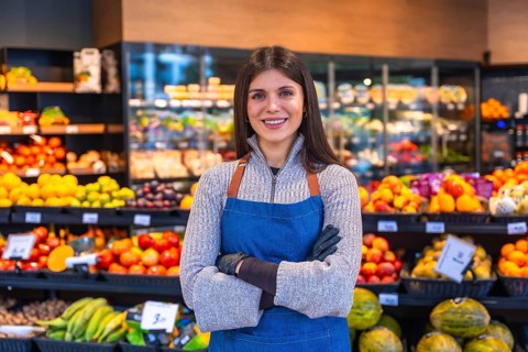 Young woman working in a fruit and vegetable section of a grocery store, wearing an apron and