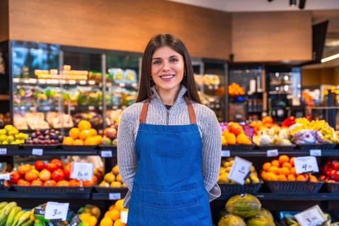 Woman wearing apron standing in a grocery store aisle, smiling at the camera, surrounded by fresh