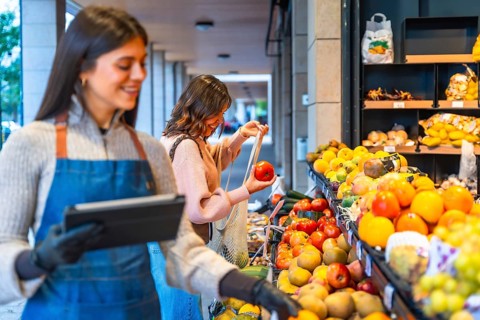 Greengrocer smiling and holding a digital tablet while a happy woman consumer chooses fresh