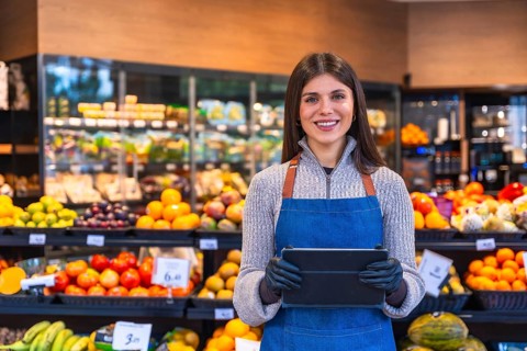 Smiling woman wearing an apron and gloves handling a digital tablet in a grocery store,