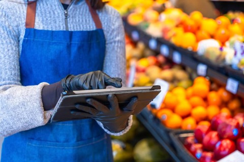 Grocery store worker wearing protective gloves and apron, using a digital tablet to manage fruit
