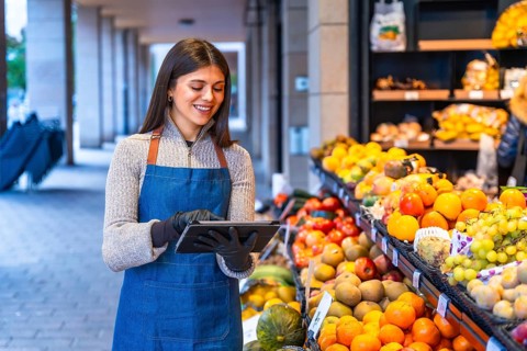 Smiling woman grocer in apron and gloves uses a tablet to manage inventory and orders at a fresh