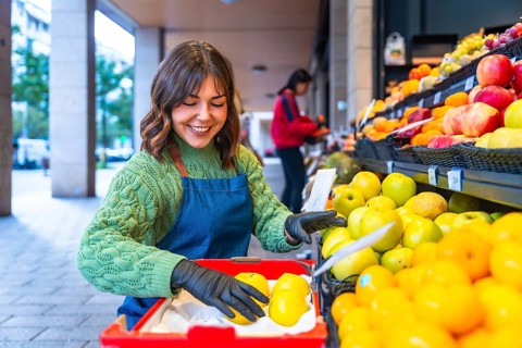 Smiling market worker in apron and gloves arranging fresh apples on a colorful outdoor fruit stall