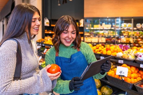 Store employee in an apron and gloves using a tablet while helping a smiling customer holding a