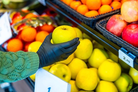 Hand in a black protective glove selects a fresh yellow apple from a supermarket produce display,