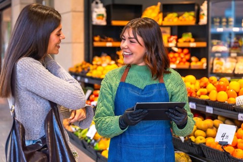 Friendly store employee wearing an apron and gloves holding a digital tablet, helping a smiling