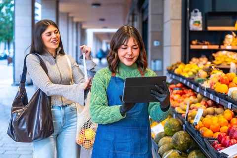 Greengrocer in apron and gloves uses a tablet to manage inventory and assist a smiling customer