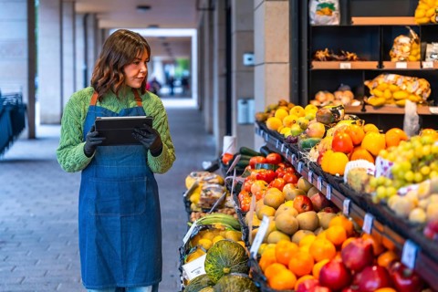 Woman grocer using a digital tablet to manage inventory and fresh produce at her outdoor fruit and