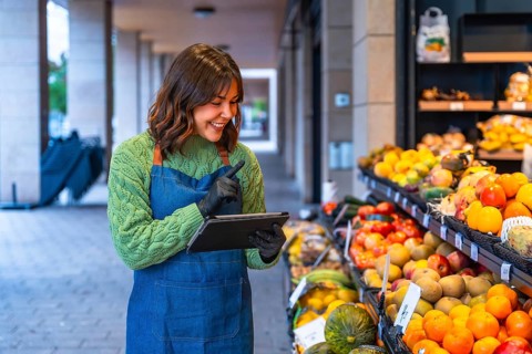 Woman in an apron and gloves managing fresh produce at her small business, using a digital tablet