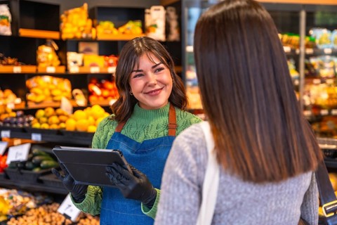 Friendly saleswoman holding a tablet and wearing gloves, assisting a customer with produce in a