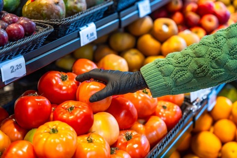 Person's hand wearing a black disposable glove picking ripe tomatoes from a display bin, promoting