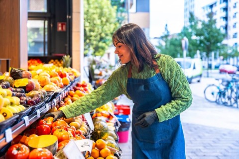 Smiling young greengrocer woman wearing an apron and gloves arranging fresh fruit and vegetables on