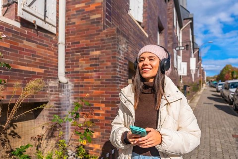 Smiling young woman walking through a sunny urban street in autumn, listening to music on