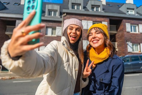 Two cheerful young women friends capturing a fun selfie on a city street, wearing winter clothing