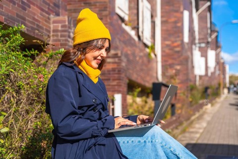 Smiling woman wearing a yellow beanie and blue trench coat, typing on a laptop while sitting