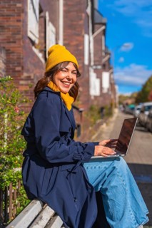 Young woman smiling at camera while sitting on a public bench in an autumn street, typing on her