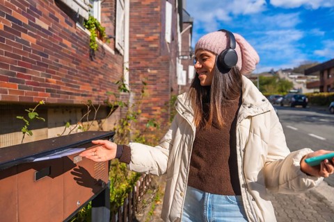 Young woman smiling, wearing a beanie and headphones, mailing a letter into an outdoor mailbox on