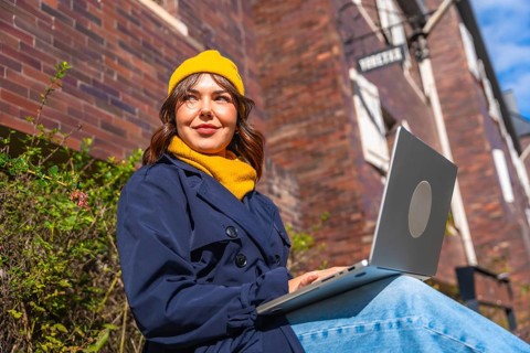 Woman wearing a warm yellow beanie and trench coat, smiling while sitting outdoors with a laptop on