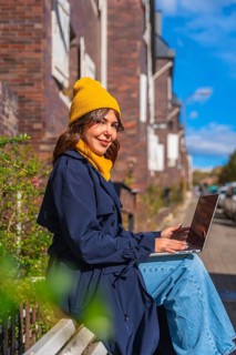 Young woman smiling while sitting on a bench, typing on her laptop, conducting remote work or