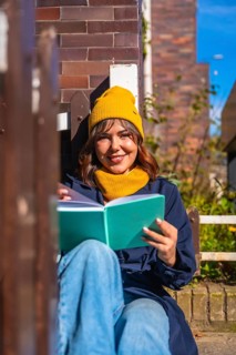Young woman in a yellow beanie sits on a bench against a brick wall, smiling and reading a book