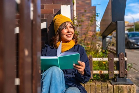Woman in casual autumn attire reading a notebook while sitting outdoors, smiling and contemplating