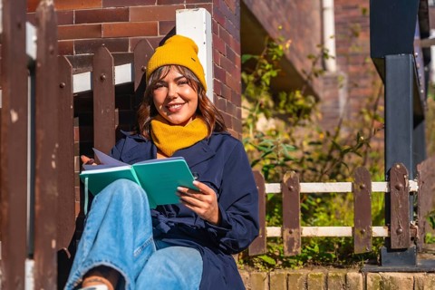 Young woman wearing a yellow beanie and scarf. Sitting outdoors in autumn sunlight. Smiling while