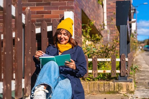 Woman wearing a yellow beanie and blue coat sitting on steps, enjoying a book in the warm sunlight