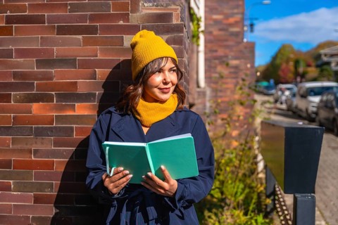 Young woman wearing a yellow beanie and scarf, holding an open journal while standing against a