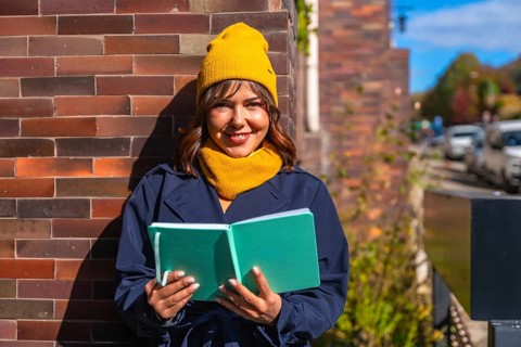 Young woman wearing a yellow beanie and scarf, dressed in a trench coat, standing outdoors in warm