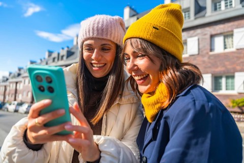 Two young women, dressed in winter hats and warm clothing, are laughing while looking at a mobile
