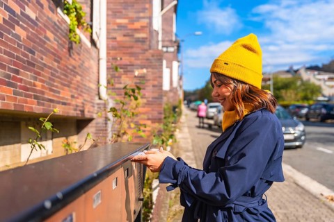 Woman wearing a yellow beanie and blue trench coat, smiling while sending a letter through a street