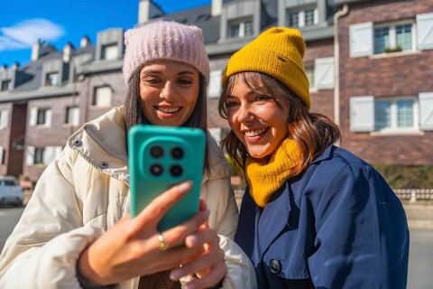 Two friends in winter jackets and beanies smiling and taking a selfie on a sunny city street,