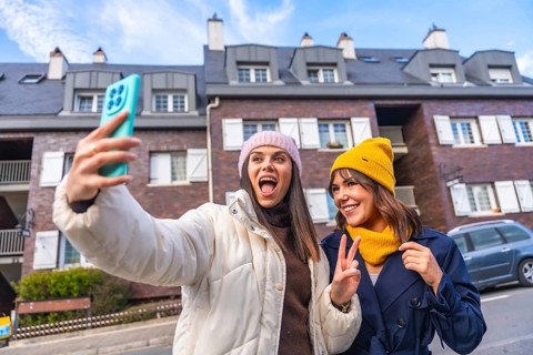 Two cheerful young women take a sunny winter selfie on a city street, bundled in beanies and