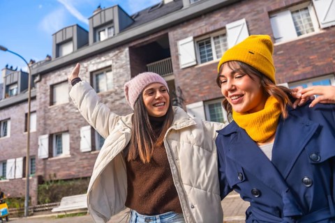 Two young women friends walking a city street in winter coats and beanies, laughing and chatting