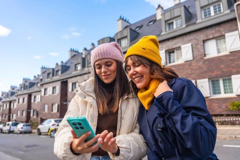 Two young women in winter coats and beanies smile and share a phone while walking a city street,