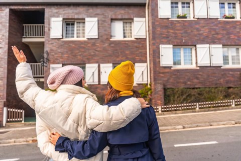 Two young women walking arm in arm down a city sidewalk in cool weather, wearing parkas and