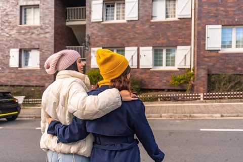 Two young women in warm winter clothes walk arm in arm down a city street, smiling and enjoying a
