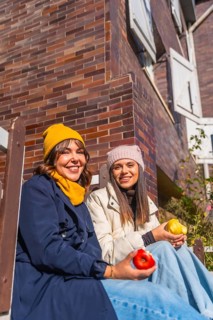 Two friends in warm winter clothes sit against a brick wall, smiling and sharing red and yellow
