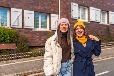 Two smiling young women friends are standing together on an urban street in front of a brick