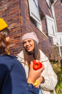 Two young women chatting outdoors on a sunny urban street, smiling and sharing apples while bundled