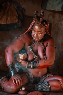 Natural perfume, elderly Himba woman smokes her hair for good smell, in a hut, traditional Himba