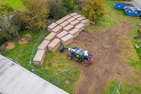 Aerial view of boxes and a forklift truck on a fenced grassy area with containers, PV