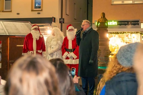 Two Santa Clauses stand with a woman and a speaker in front of an audience at a Christmas market,