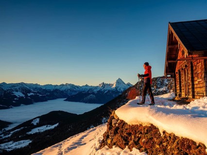 Mountaineers enjoy the sunrise in front of an alpine hut in winter, the Watzmann behind, fog in the