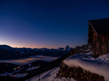 Alpine panorama in winter with Watzmann and alpine hut under starry sky at dawn, fog in the valley,