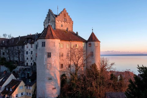 The historic Meersburg Castle, illuminated by the setting sun, right behind it Lake Constance with