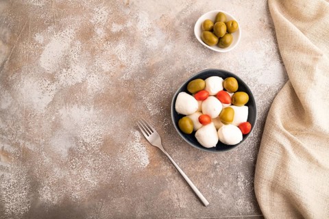 White Mozzarella cheese, with tomatoes and olives in blue bowl on brown concrete background and